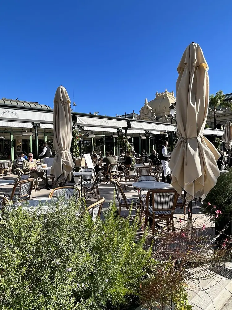 The open terrace of the Cafe de Paris in Monte-Carlo, rattan chairs and marble tables occupied by people in everyday clothes, with the ornate rooflines of Place du Casino rising behind the awnings