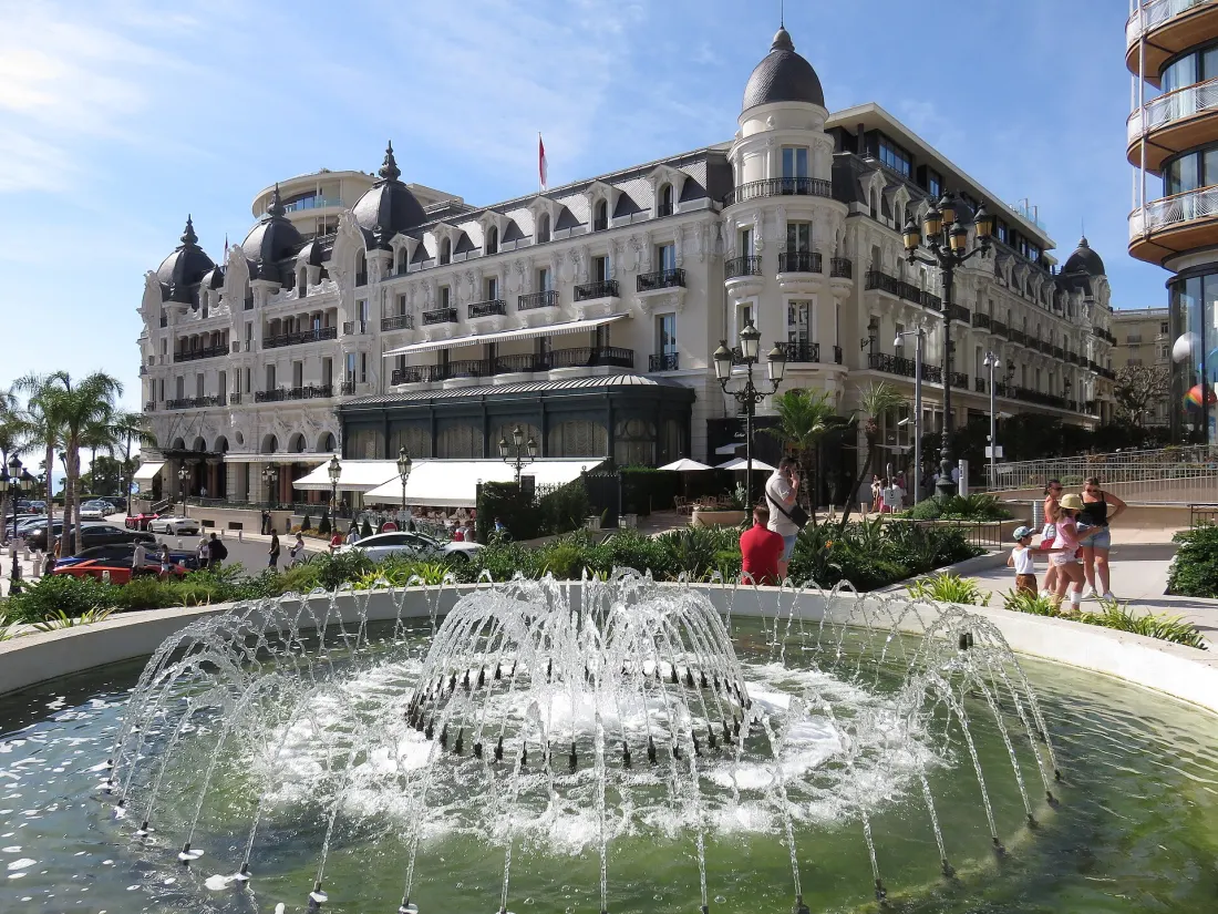 The Hotel de Paris Monte-Carlo across the fountain on Place du Casino, its slate domes and awnings above the terrace, with tourists in shorts and t-shirts standing on the pavement outside