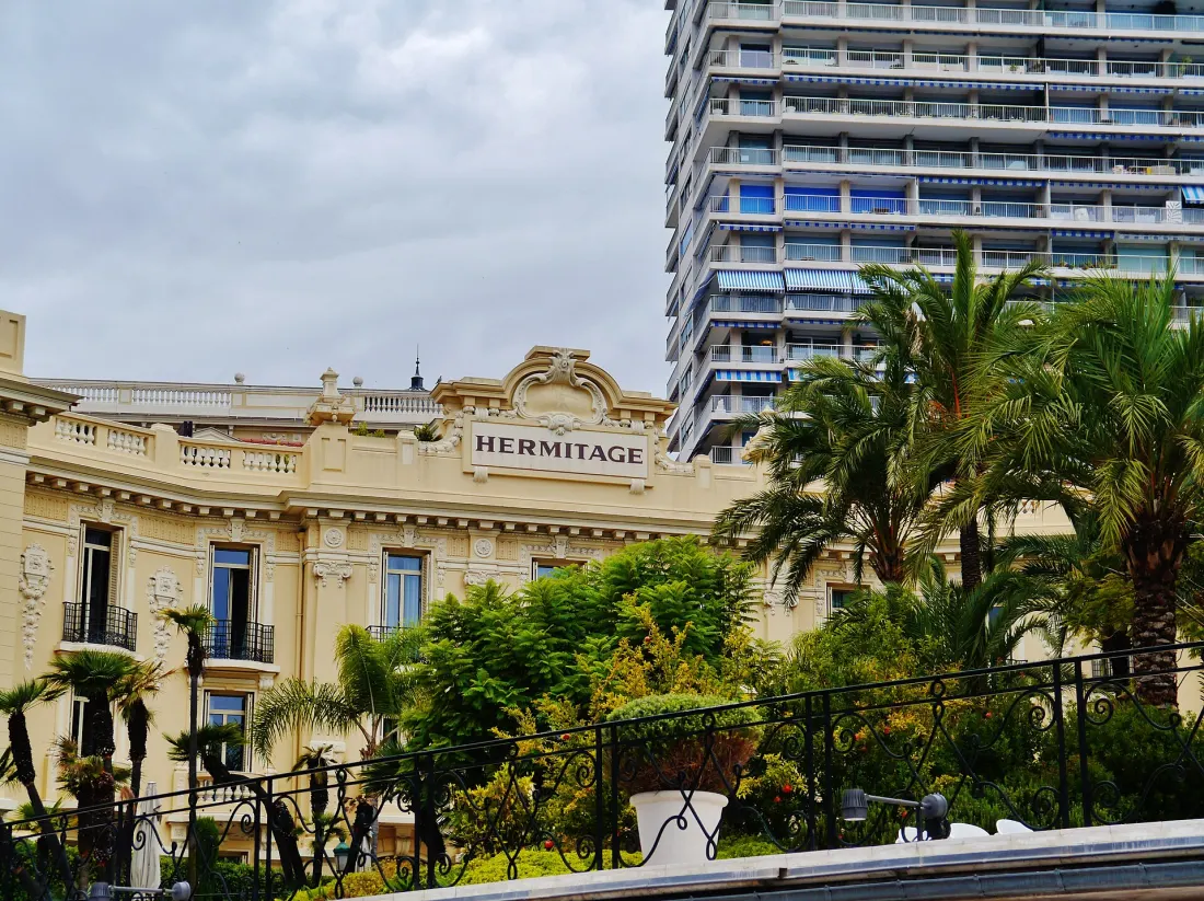 The cream Belle Epoque frontage of the Hotel Hermitage with its name carved above the cornice, palms and wrought iron railings in front, and a tall modern residential tower standing directly behind it