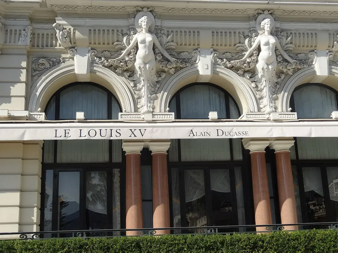 The Le Louis XV Alain Ducasse awning on the Hotel de Paris, set beneath arched windows and two carved caryatid figures in the Belle Epoque stonework