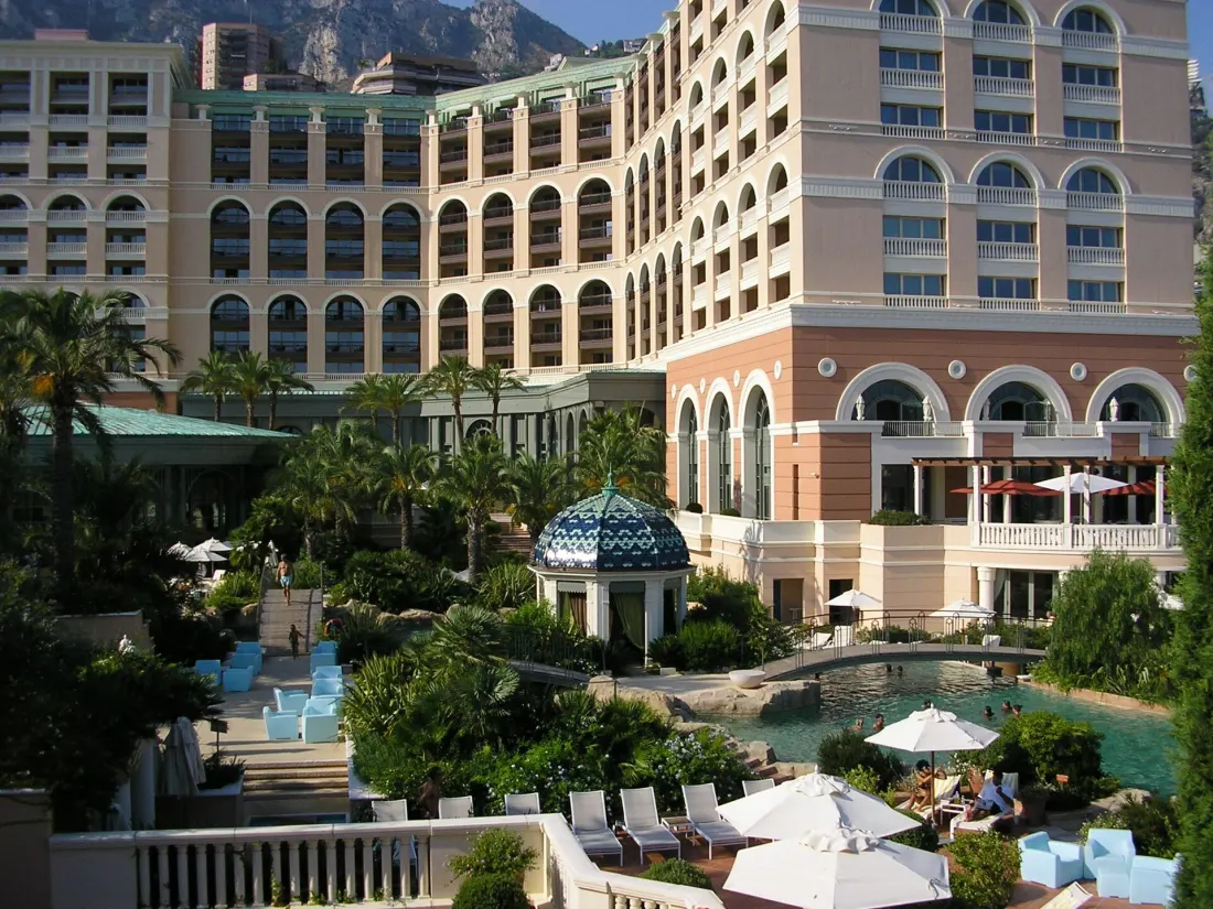 The Monte-Carlo Bay Hotel and Resort seen from inside its grounds, a large arcaded block rising above the sand-bottomed lagoon, with loungers, parasols and a tiled gazebo in front of it